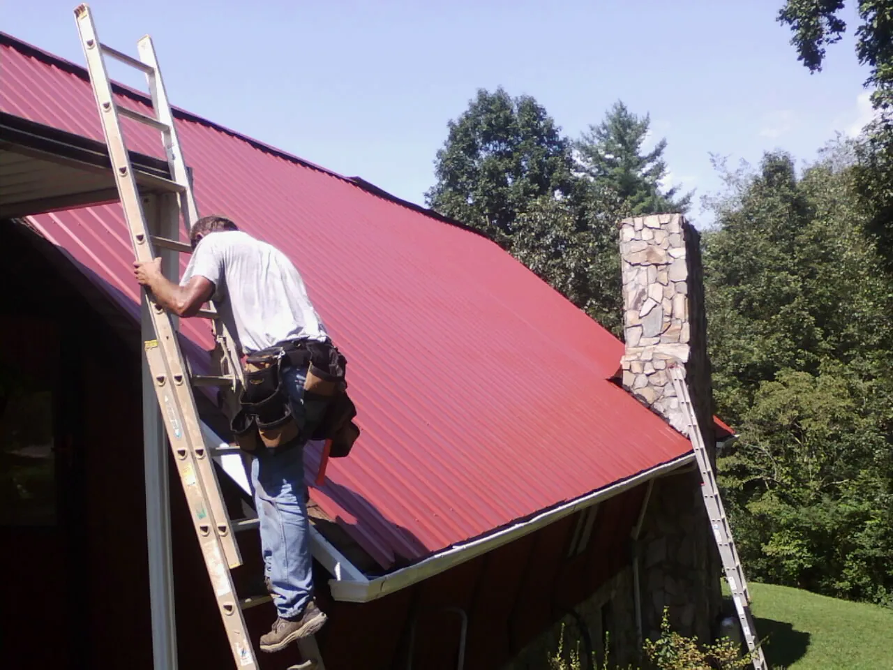 Roofer on a ladder beside a red metal roof and stone chimney — Soffit & Fascia Repair by Roofing Brothers in Berea